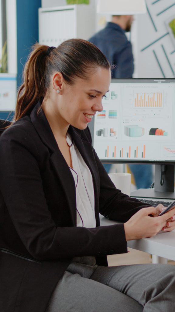 corporate-employee-looking-smartphone-screen-while-working-business-planning-with-computer-charts-company-office-businesswoman-using-mobile-phone-brainstorming-ideas-min