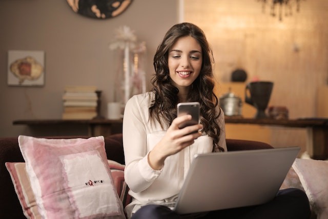 A woman sitting on the bed with a phone in her hand and a laptop in her lap